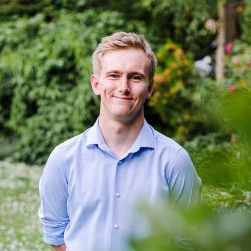 Medium shot of Toby Jones (blond male) smiling, wearing a light blue shirt standing amongst greenery 