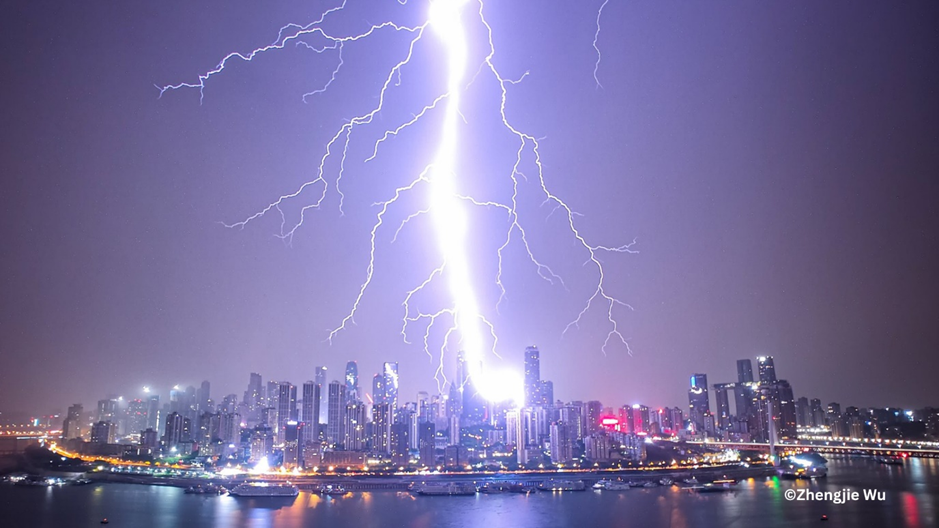 Photo of an extremely bright lightning bolt striking a coastal city at night. The sky is illuminated purple by the flash. Photo by Zhengjie Wu.