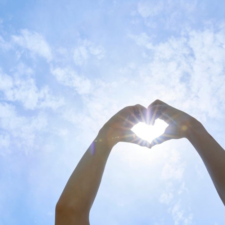 A pair of hands form a heart shape in front of the sky